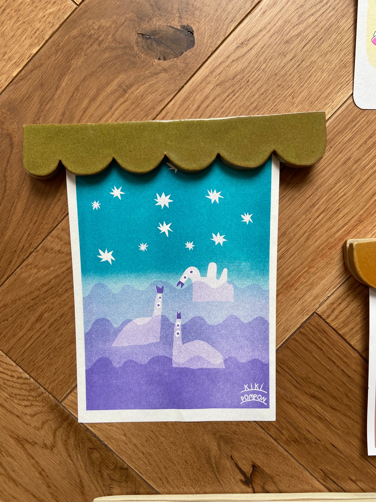 Porte affiche vert foncé tenant une carte de Kiki Pompon représentant des cignes dans le ciel