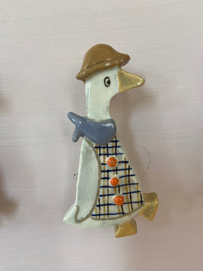 Patere représentant une oie avec un chapeau marron. L'oie à un foulard bleu et une chemise 